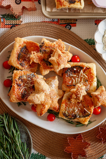 Crispy Parmesan and Broccoli Puff Pastry Appetizers on a white platter with tomatoes as a garnish. The Platter is on a rattan placement which has a holiday tea towel with trucks and Christmas trees. There is a plate with rosemary on it and a bowl with grated Parmesan to the side.