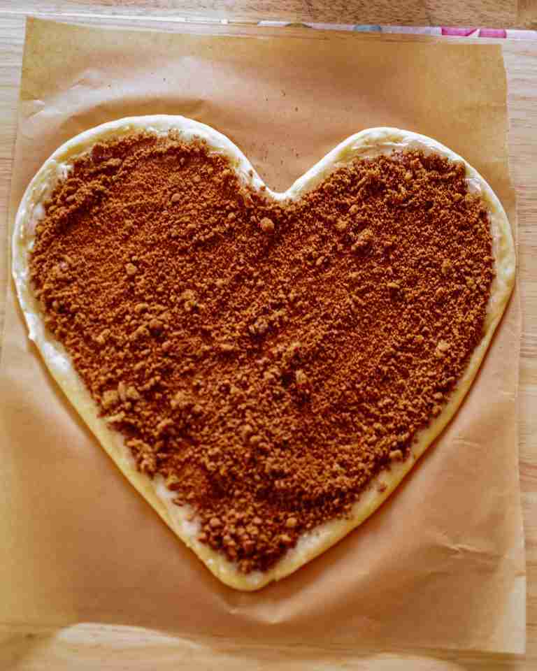 20240104-DSC_0212 (1) Heart shaped cinnamon roll dough with brown sugar and cinnamon spread evenly over it, on a piece of brown parchment paper.