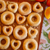 Orange buttermilk donuts on parchment paper on a wooden board with a floral tea towel in the background and three oranges in the top right corner.