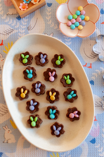 Fifteen pieces of fudge shaped like flowers with m&ms on them making a bunny shape. The fidge is on a white ceramic tray and there is a flower dish int he background with pastel m&ms on it. There is a light blue tea towel in the background.