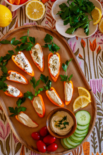 Spring veggie board with nine halved mini orange peppers that re stuffed with cream cheese or hummus and have parsley coming out of the tops to make them look like carrots. There is a dish of hummus on the board, sliced cucumbers, grape tomatoes and two lemon slices.