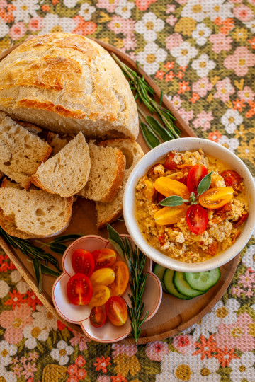 A wooden tray with half a loaf of bread, several smaller cut pieces of bread and a bowl of roasted tomatoes and feta with fresh tomatoes and sage on top. There is a flower shaped dish with sliced tomatoes on it and rosemary, sage and some sliced cucumbers as garnish on the tray.