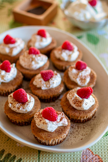 Ten mini cappuccino flavored cheesecakes with whipped cream and a raspberry on top. They are on a white tray with a green tea towel in the background.