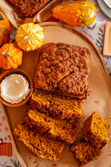 Pumpkin bread with a cream cheese swirl and a cinnamon streusel topping on a wooden serving board with a small container of cream cheese and gourds in the background.