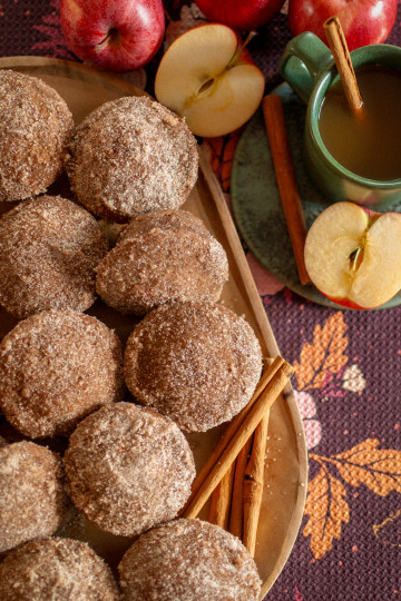 Apple cider donut muffins on a wooden tray with a mug of apple cider and cinnamon sticks in the background.