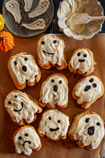 Nine ghost shaped cinnamon rolls with cream cheese frosting and faces made with black cocoa powdered sugar.