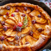 Deep dish apple galette with frangipane and sugared cranberries in a cast iron skillet, with a holly tea towel in the background.