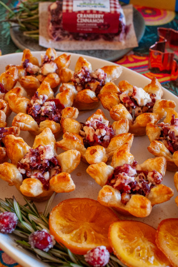 Cranberry cinnamon goat cheese tarts made with puff pastry and homemade cranberry sauce. They are on a white tray, garnished with candied oranges, rosemary and sugared cranberries.