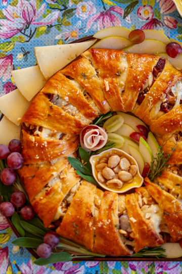 Ham and cheese wreath with roasted grapes and pears on a round wooden tray, garnished with fresh fruit.
