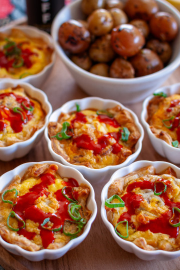 Six mini quiches in scalloped ramekins topped with green onions and hot sauce, on a wooden serving board. There is a bowl of mini potatoes on the board as well.