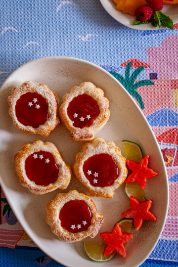 Five puff pastry seashells with a guava filling and three star sprinkles on each. They are on a white ceramic tray with three watermelon starfish cutouts and four lime slices.