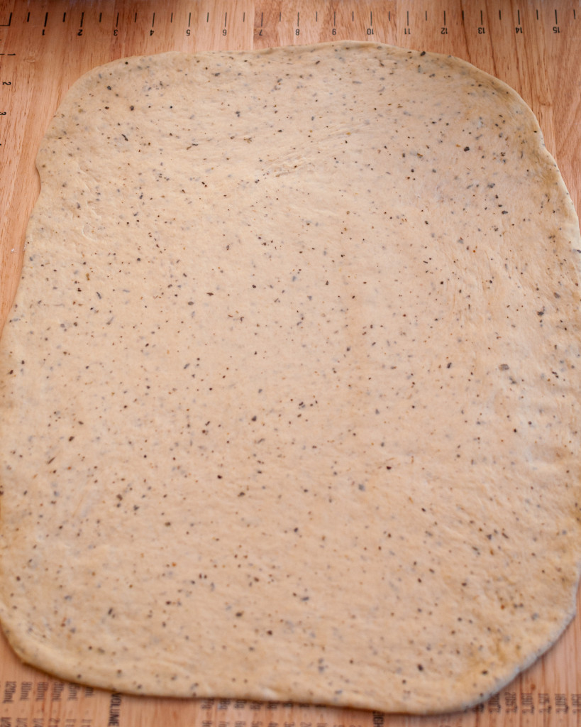 Seasoned pizza dough rolled out into a rectangle on a wooden pastry board.
